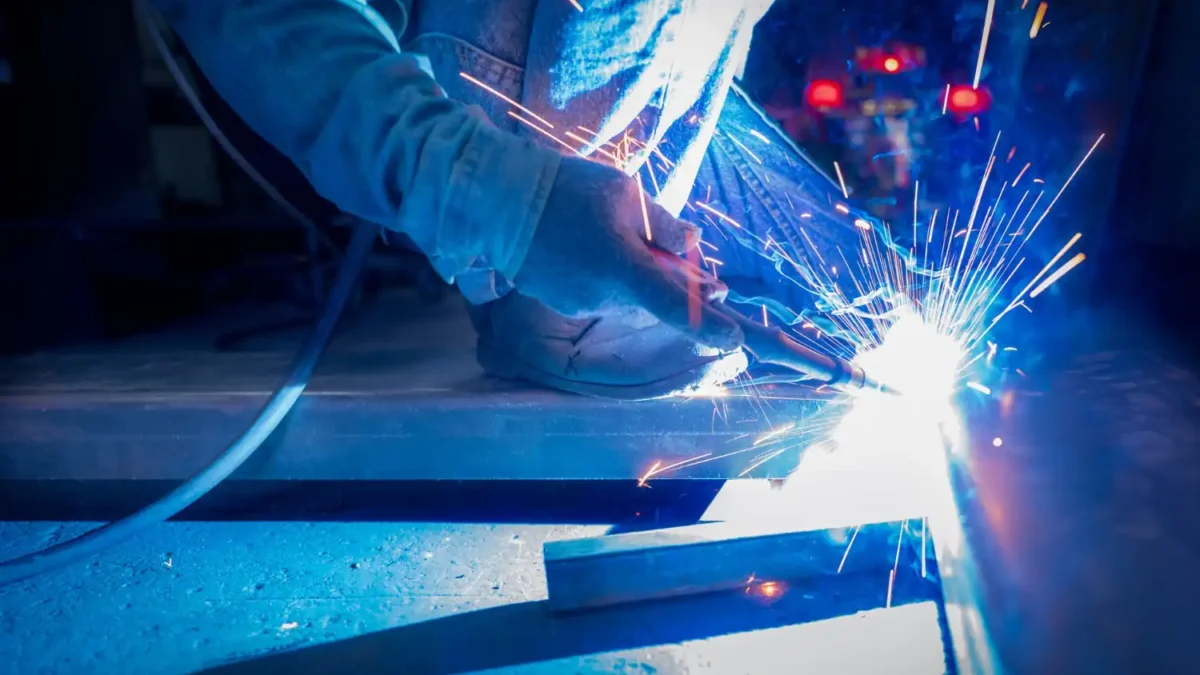 Trabalhador realizando soldagem em metal, com faíscas azuis e laranjas se espalhando durante o processo.