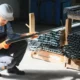 Trabalhador com capacete e luvas de proteção inspecionando barra metálica em depósito de tubos e perfis de aço