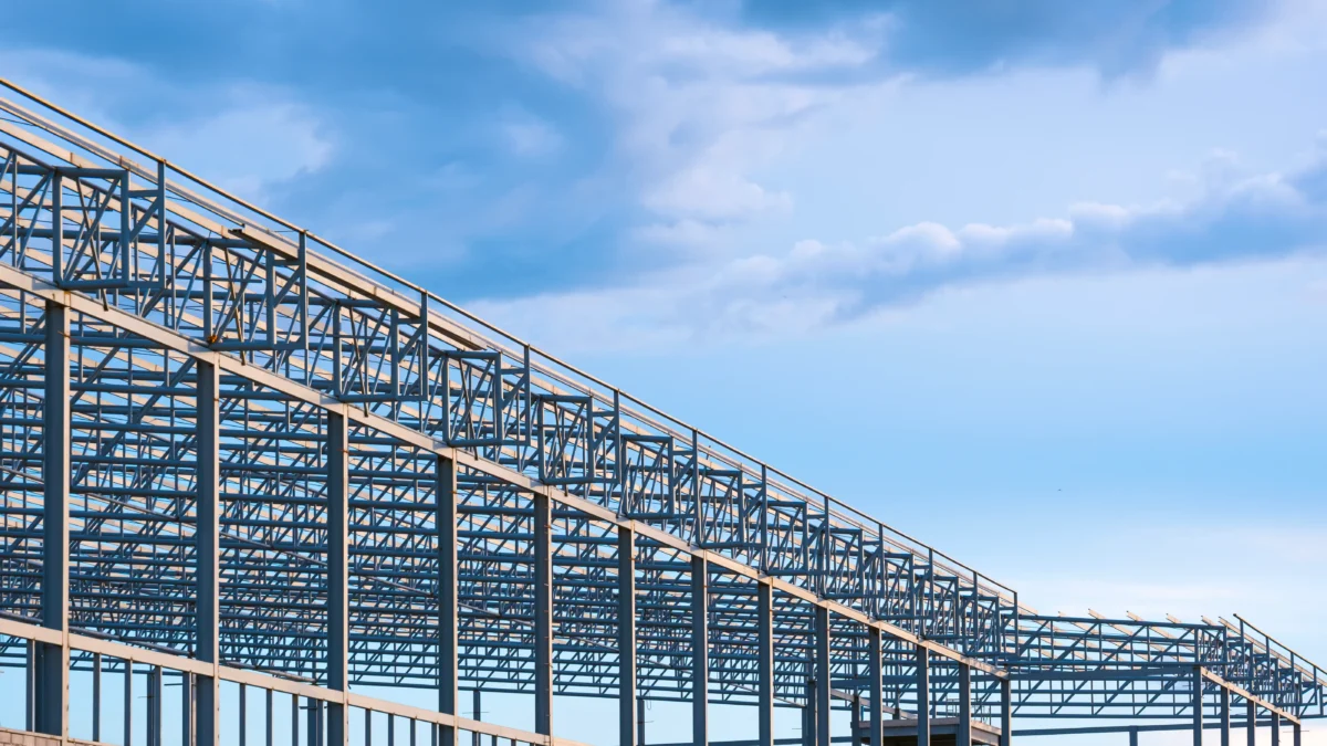 Estrutura metálica de galpão industrial em construção contra o céu azul com nuvens.