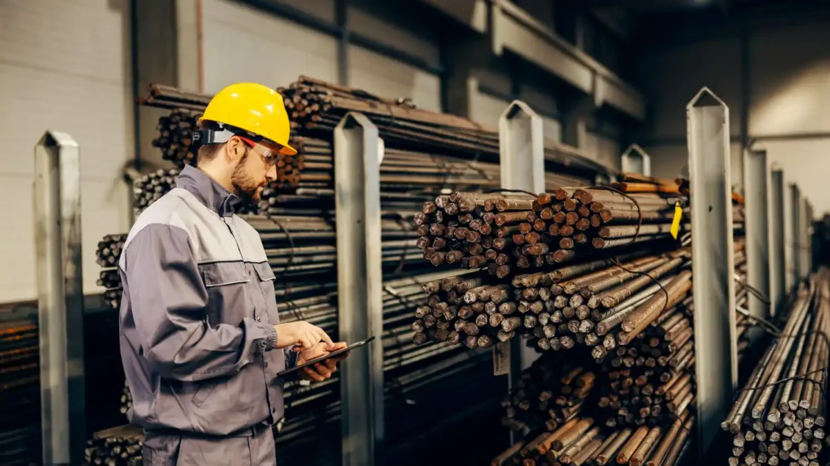 Trabalhador da indústria metalúrgica inspecionando barras de aço em um depósito, simbolizando a produção moderna de aço inoxidável.